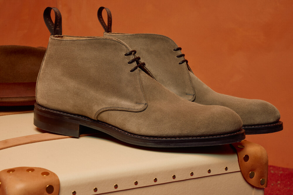 Sand-coloured chukka boots sat on a suitcase on an orange background.
