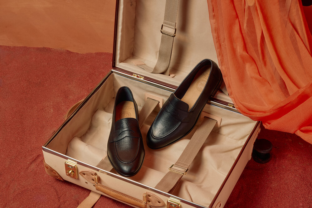 Black leather penny loafers on a suitcase on an orange background.