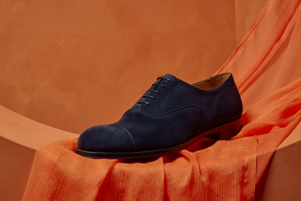 Singular blue suede Oxford shoe on an orange background.